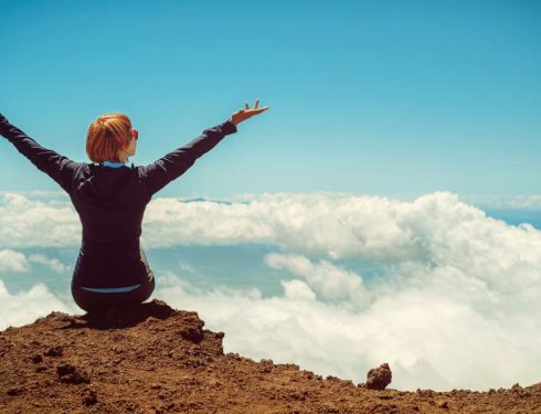 A woman enjoys a scenic view atop a cliff in Kula, Hawaii, surrounded by clouds and blue sky.